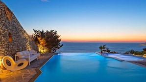 A poolside area with a stone wall, a lounge chair, and a view of the sea at sunset. - Apanema Aegean Luxury Hotel (Mykonos)