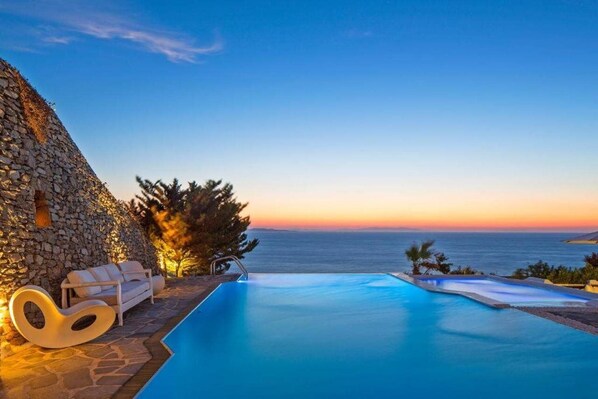 A poolside area with a stone wall, a lounge chair, and a view of the sea at sunset.