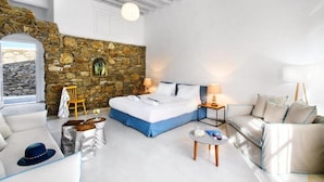 A modern room with a stone wall, a bed, a sofa, and a small table. - Apanema Aegean Luxury Hotel (Mykonos)