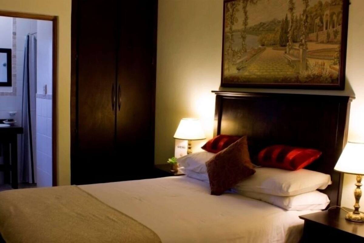 A hotel room with a bed, two bedside lamps, a painting above the bed, and a door leading to a bathroom.