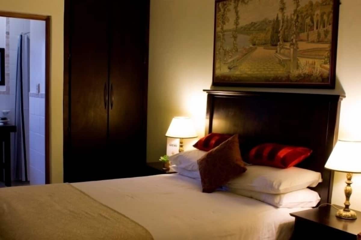 A hotel room with a bed, two bedside lamps, a painting above the bed, and a view of a bathroom with a shower and toilet.