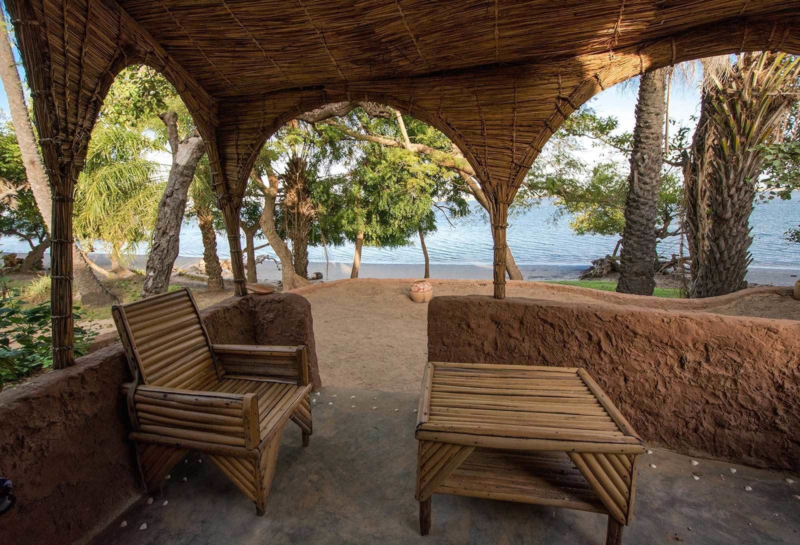 Un espacio de estar al aire libre cubierto con dos sillas de madera y una mesa, con vista a un cuerpo de agua rodeado de árboles.