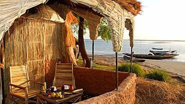 Eine strohgedeckte Hütte mit zwei Holzstühlen und einem Tisch mit Lebensmitteln, mit Blick auf einen Strand mit Booten.