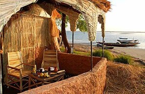 A thatched-roof hut with two wooden chairs and a small table with food items, overlooking a beach with boats. - L'Ecolodge de Simal (Fatick)