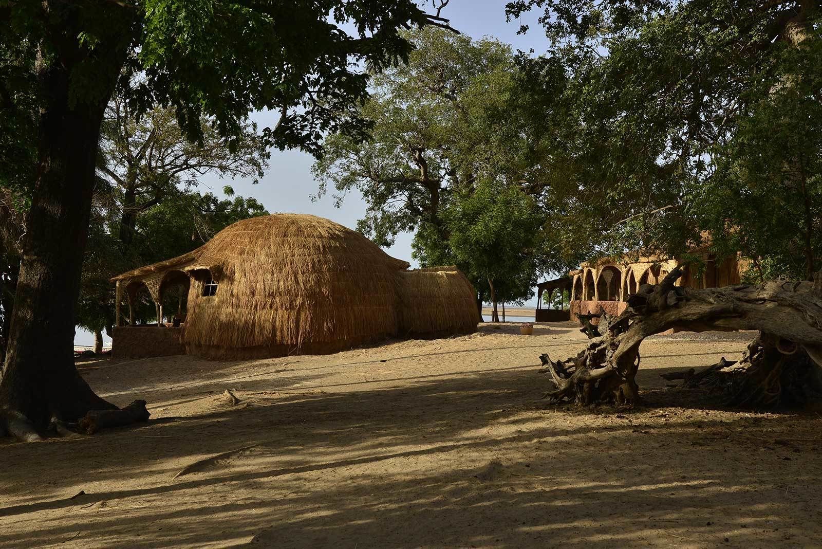 Una cabaña tradicional con techo de paja y una pequeña estructura adosada, rodeada de árboles y suelo de tierra.