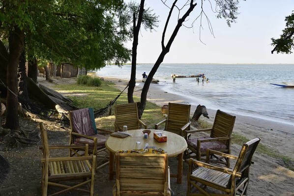 A beachside dining area with a round table and wooden chairs, overlooking a calm sea with boats and people in the background. - L'Ecolodge de Simal (Fatick)