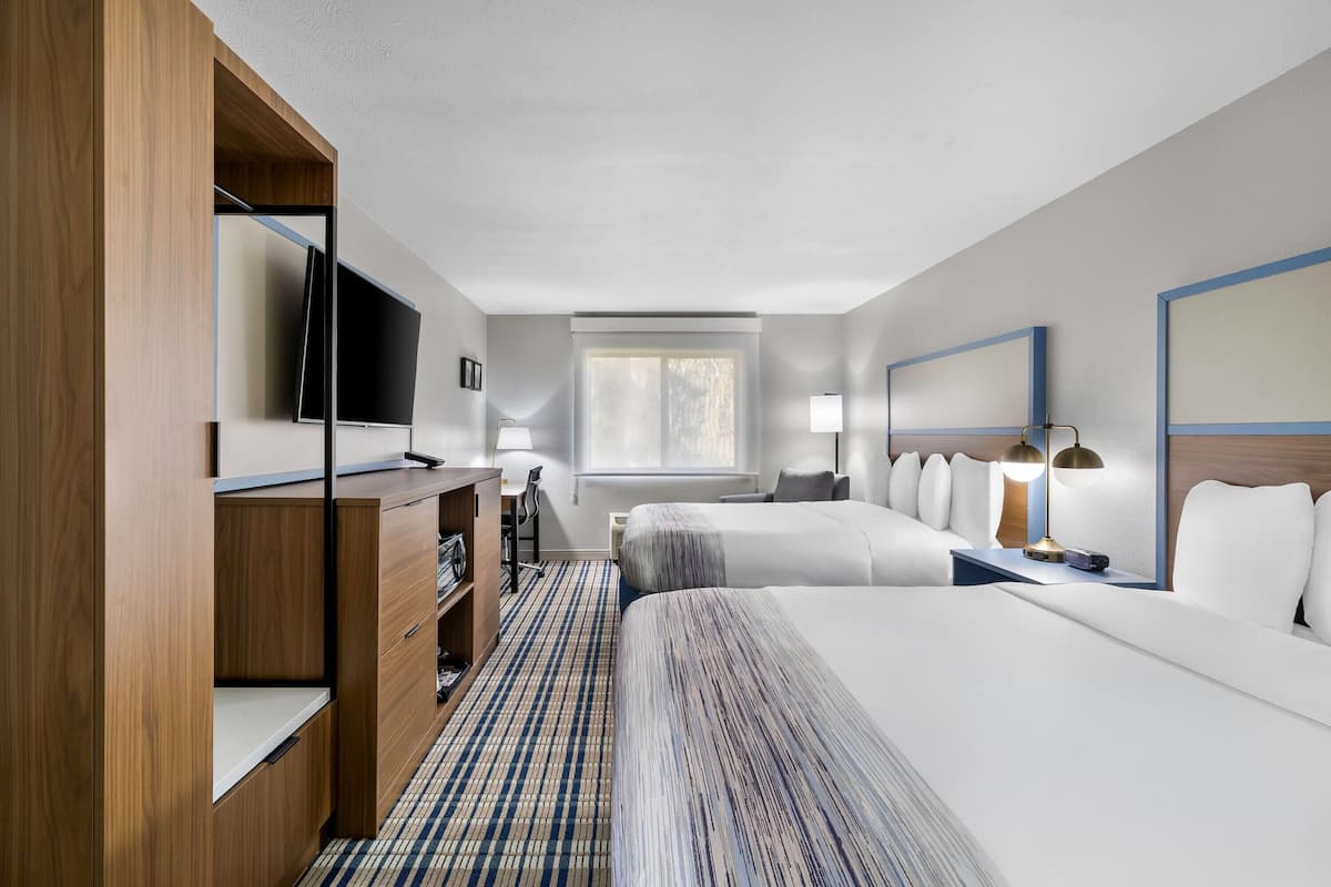 A hotel room with two beds, a desk with a TV, a chair, a lamp, and a window.