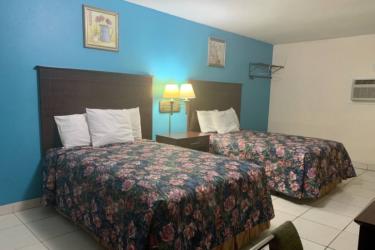 A hotel room with two beds, each with a floral bedspread, a nightstand with a lamp, and a wall-mounted shelf.