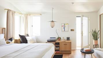 A bedroom with a large bed, bedside tables, a desk with a chair, a potted plant, and a door leading to another room.