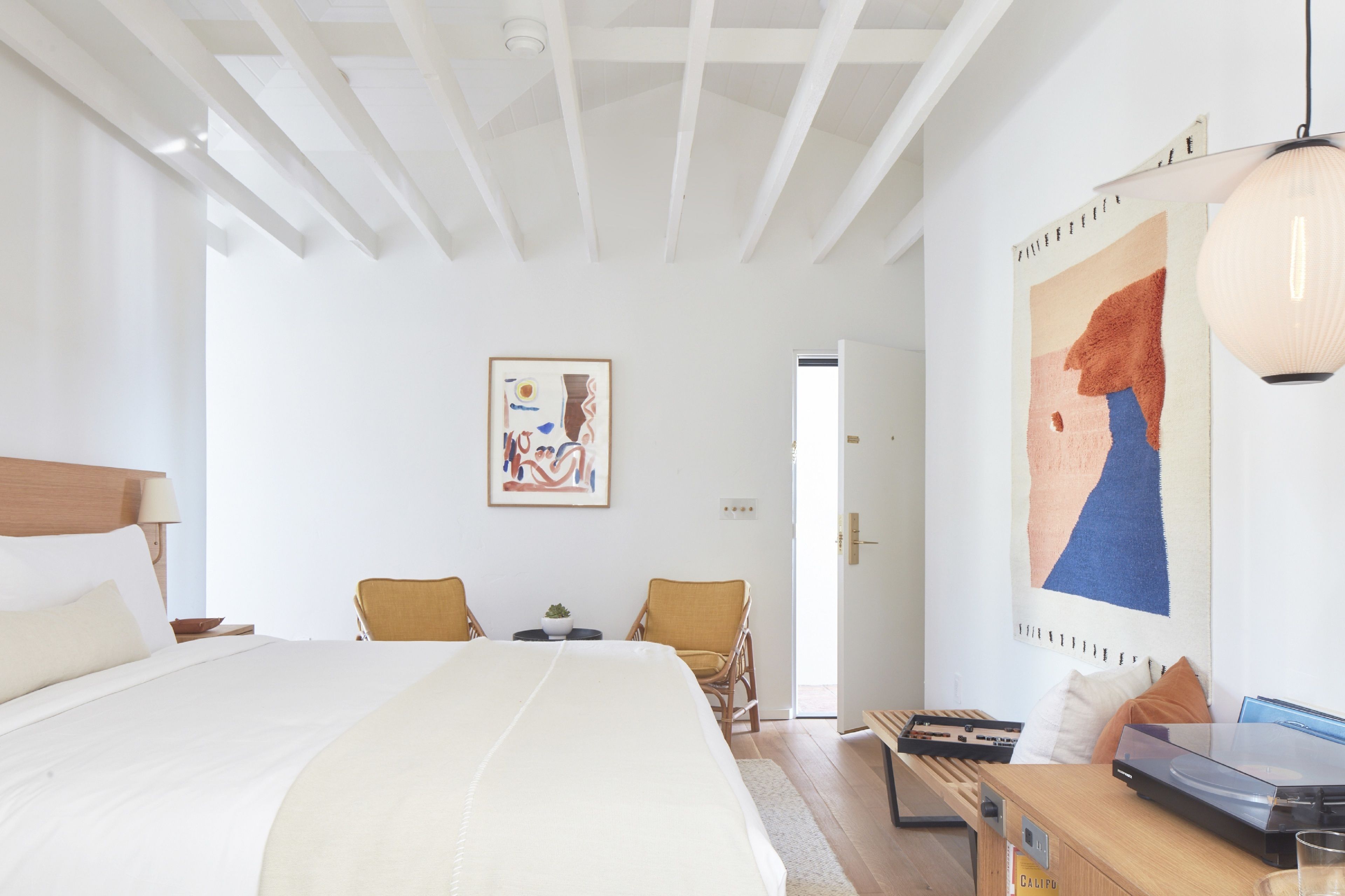 A spacious bedroom with a large bed, a desk, and a chair. There is a painting on the wall and a door leading to another room.