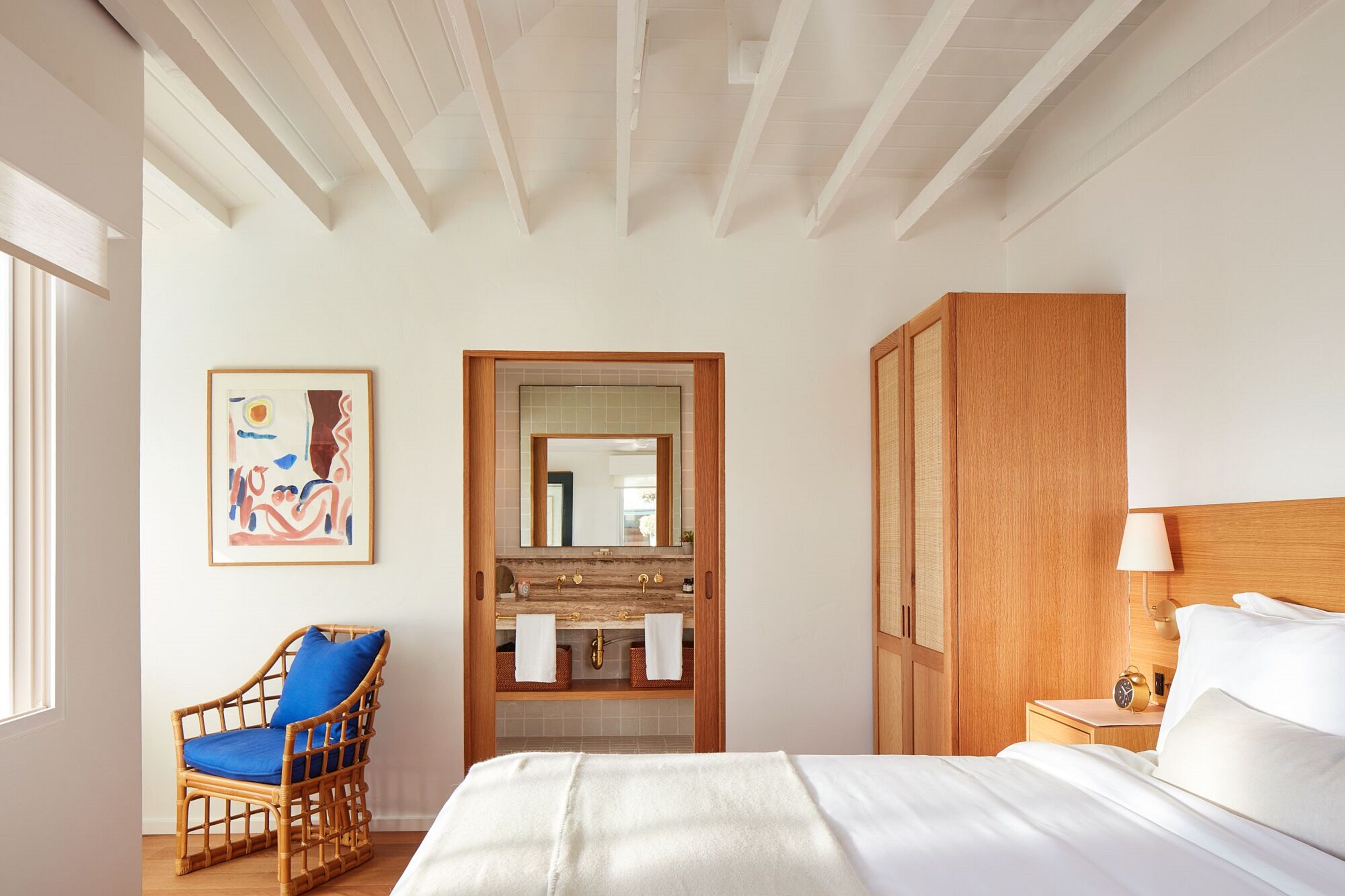 A hotel room with a bed, a wooden wardrobe, a bathroom with a mirror and sink, a wicker chair, and a painting on the wall.