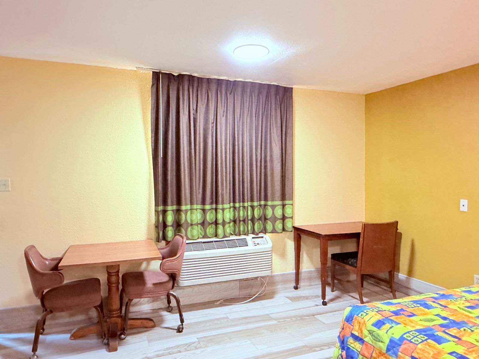 Una habitación pequeña y luminosa con una mesa de madera y dos sillas, una cama con un edredón colorido, y una ventana con cortinas.