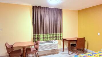 Una habitación pequeña y luminosa con una mesa de madera y dos sillas, una cama con un edredón colorido, y una ventana con cortinas.