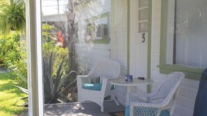 Un porche abrité avec deux fauteuils en osier et une petite table, une maison blanche aux bordures vertes, et le chiffre « 5 » peint sur le mur.