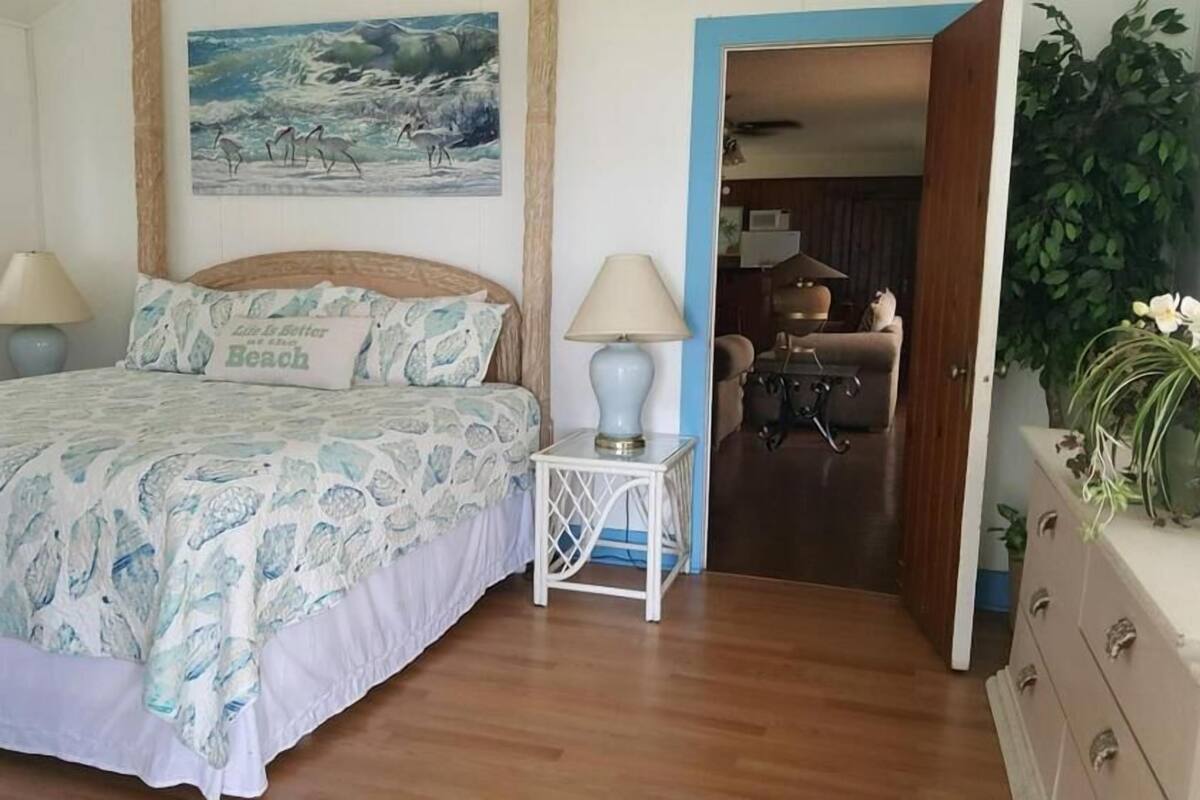 Een slaapkamer met een bed, twee bedlampen, een schilderij van een strand en uitzicht op de woonkamer.