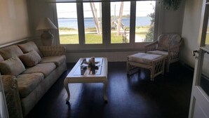Un salon chaleureux avec une grande fenêtre donnant sur un plan d’eau, un canapé en osier, une table basse en bois et un fauteuil en osier.