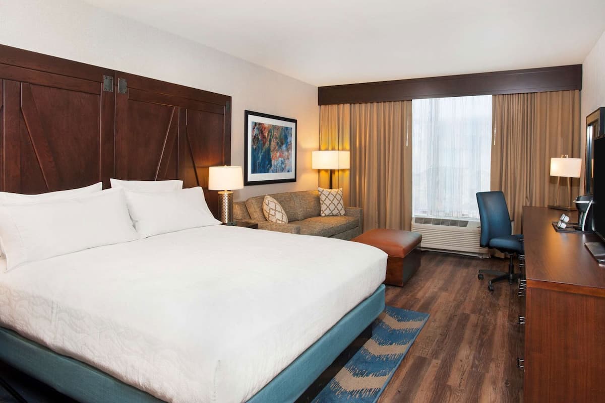 Habitación de hotel con una cama grande, un sofá, un escritorio con televisor y una ventana con cortinas.