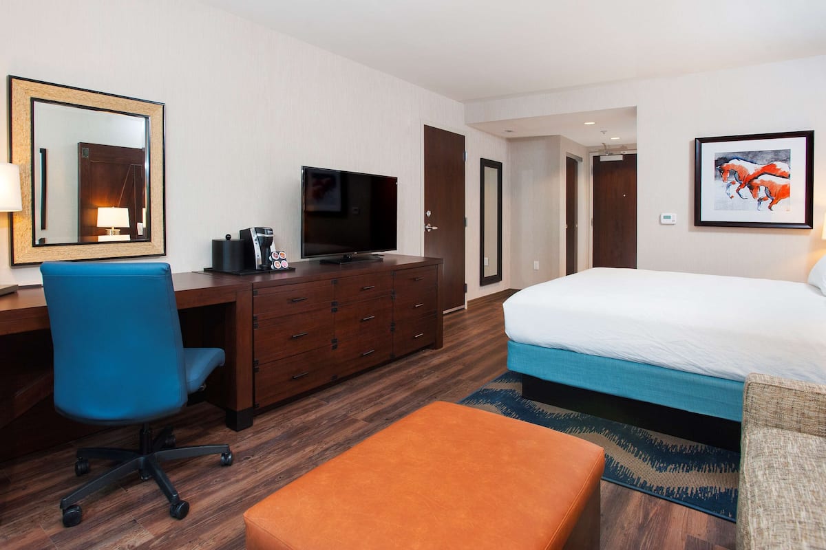 Habitación de hotel con una cama, un escritorio con televisor, una silla, cafetera y un cuadro en la pared.