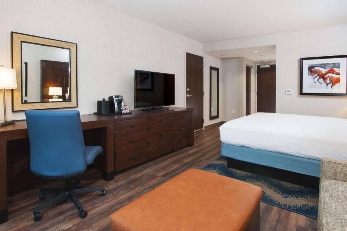 Habitación de hotel con cama, escritorio con televisor, una silla, cómoda y un cuadro en la pared.