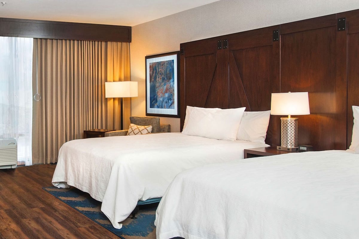 Habitación de hotel con dos camas, un cuadro grande, una ventana con cortinas y un aire acondicionado instalado en la pared.