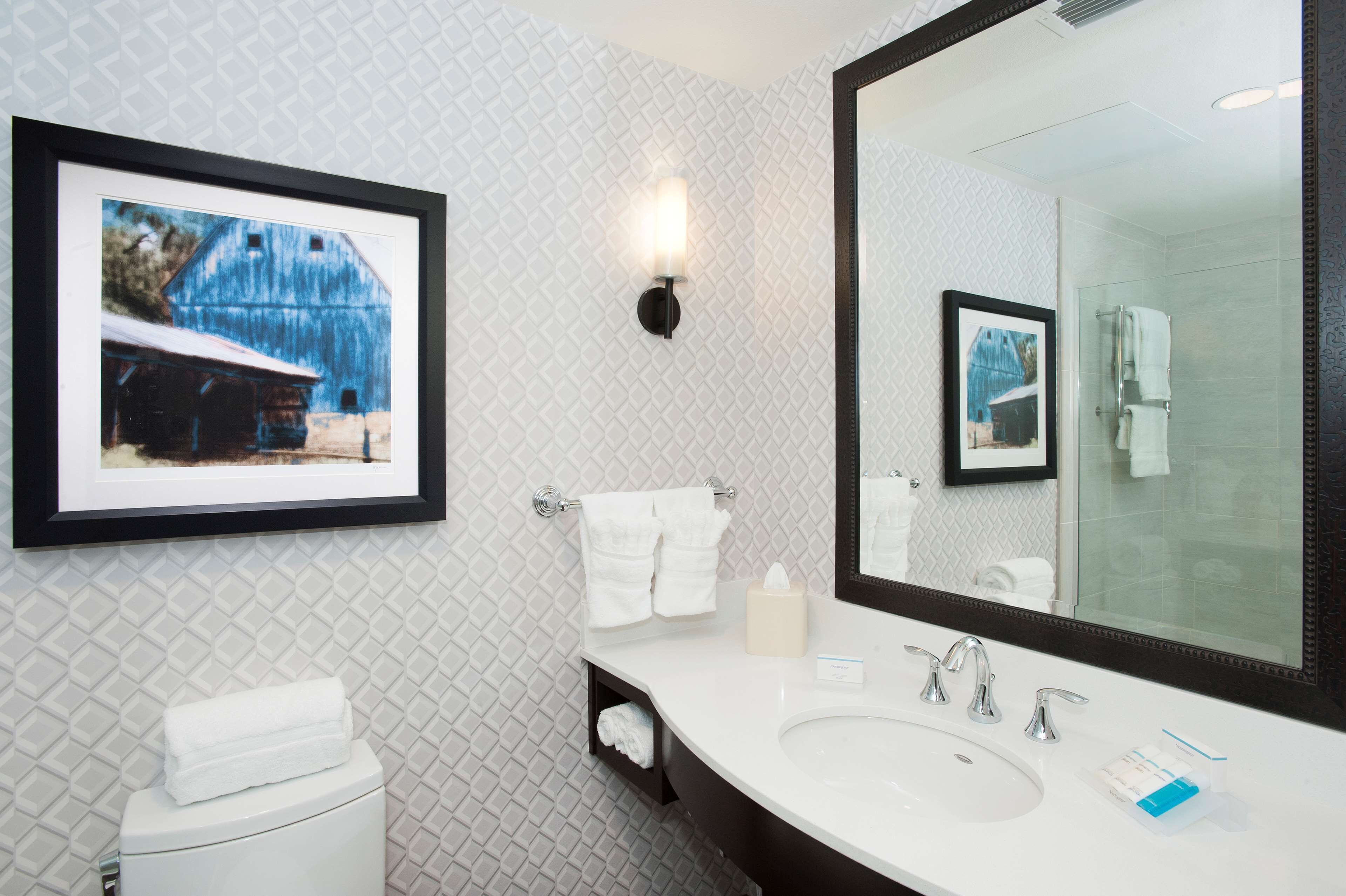 Une salle de bain avec un grand miroir, un lavabo blanc et un tableau encadré représentant une grange, accroché au mur.