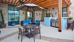 Un patio al aire libre con una mesa redonda y sillas, asientos acolchados azules, una estación de parrillada y una pérgola.