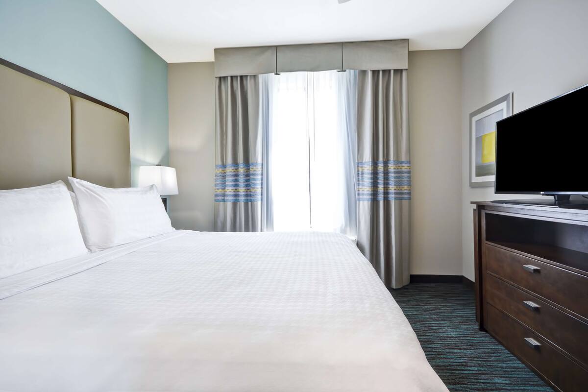 A hotel room with a large bed, a flat-screen TV, a dresser, and a window with curtains.