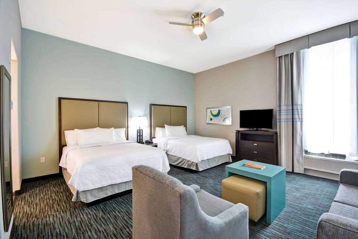 A hotel room with two beds, a desk, a chair, a television, and a ceiling fan.