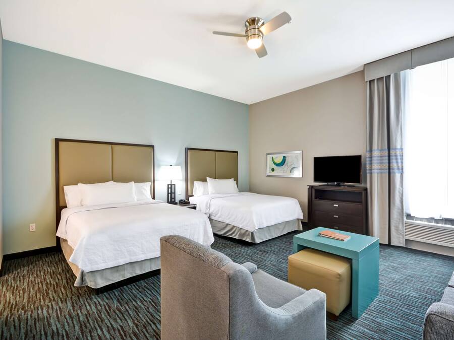 A hotel room with two beds, a desk, a chair, a television, and a ceiling fan.