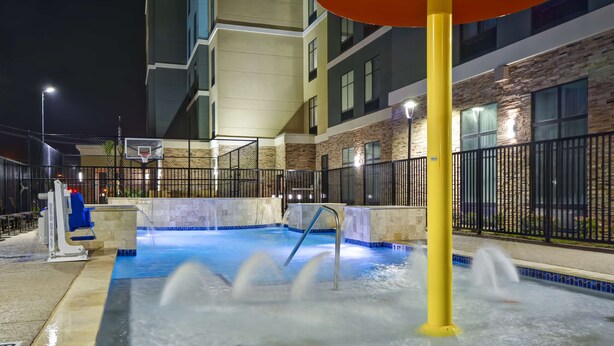 Zona de piscina exterior con canasta de baloncesto, fuente de agua y edificio de fachada de piedra con múltiples ventanas.