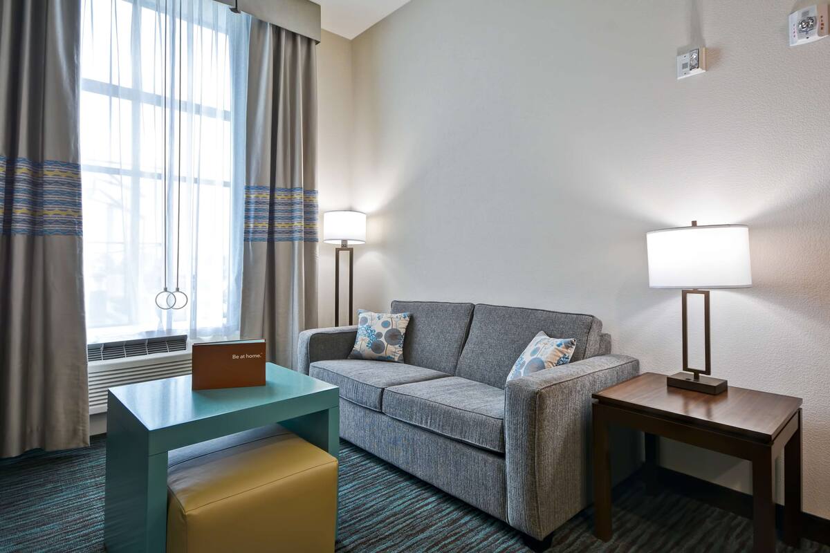A hotel room with a sofa, a small table, a lamp, and a sign on the table.