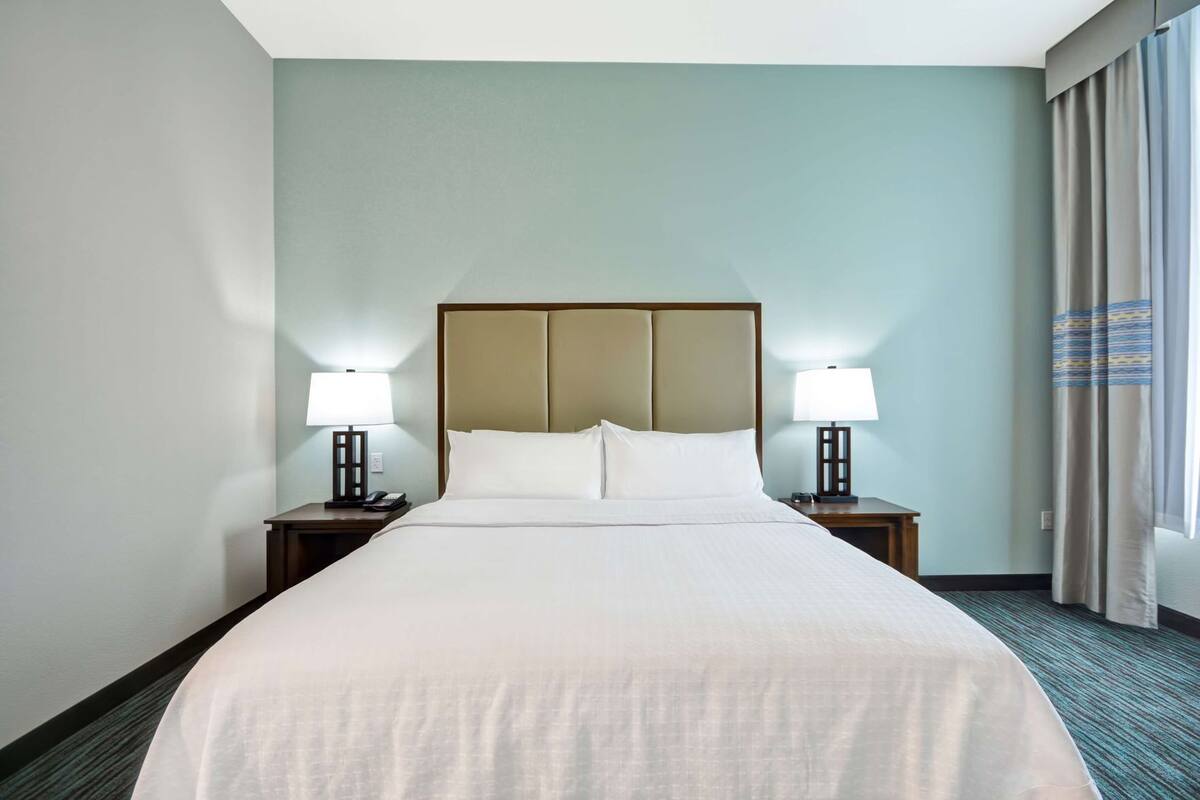 A hotel room with a large bed, two bedside tables with lamps, a headboard, and a view of the outside through a window with curtains.
