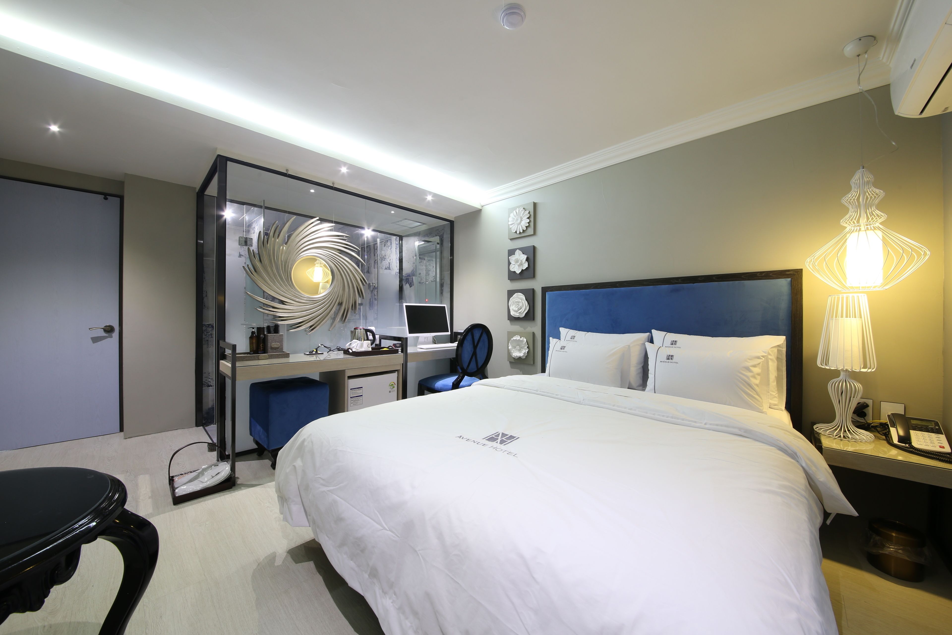 A modern hotel room with a large bed, a desk with a computer, a chair, and a bedside table with a lamp.