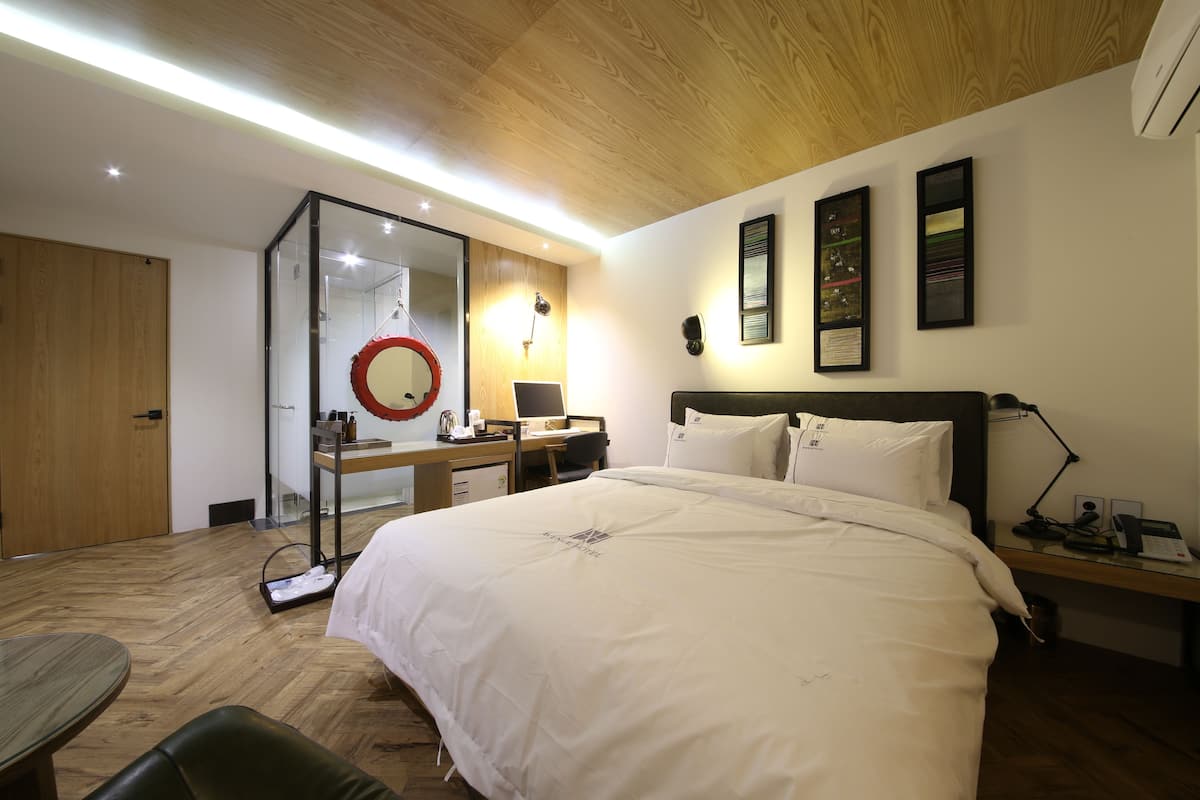 A modern hotel room with a large bed, a desk with a computer, a mirror, and a small seating area.