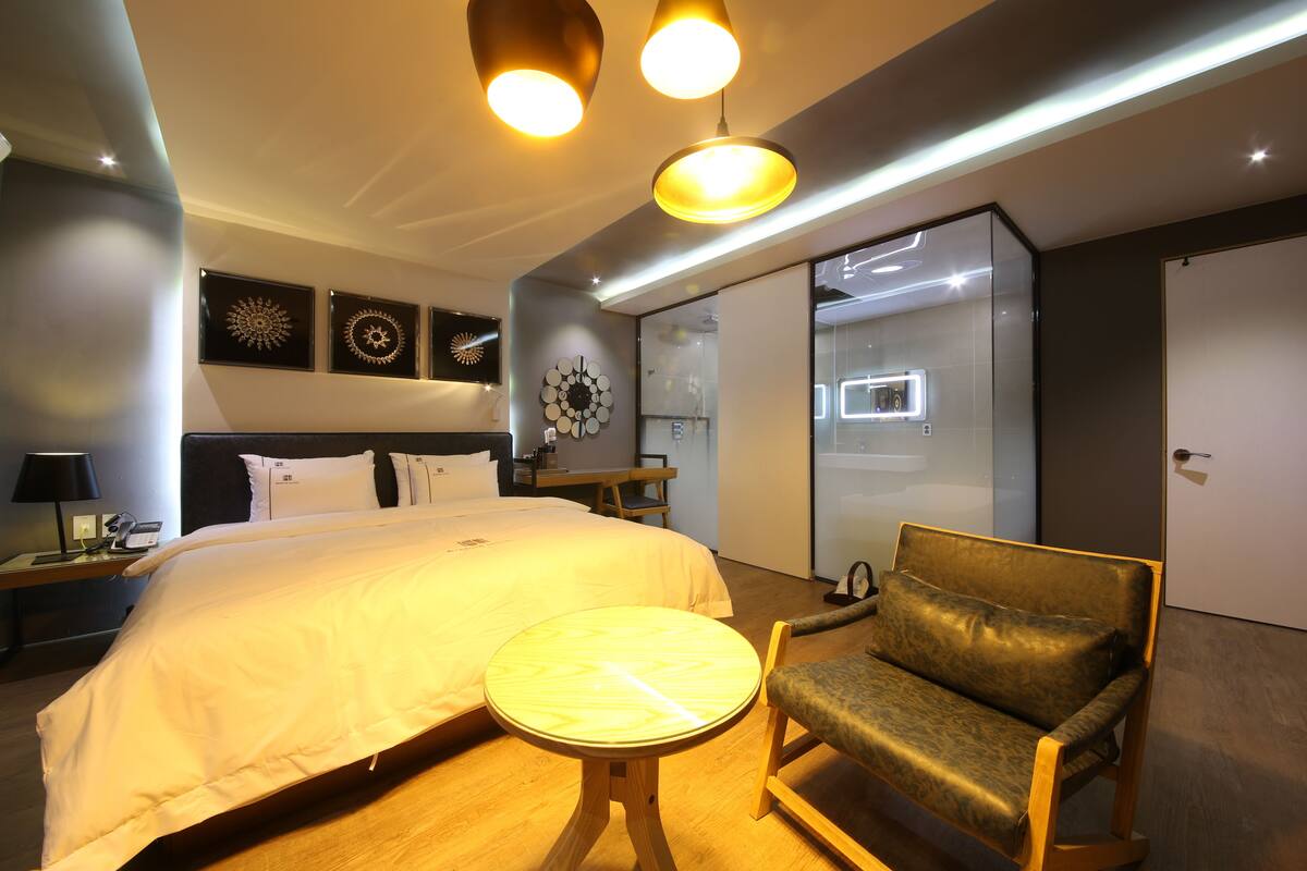 A modern hotel room with a large bed, a round table, a leather armchair, and a wall-mounted clock.