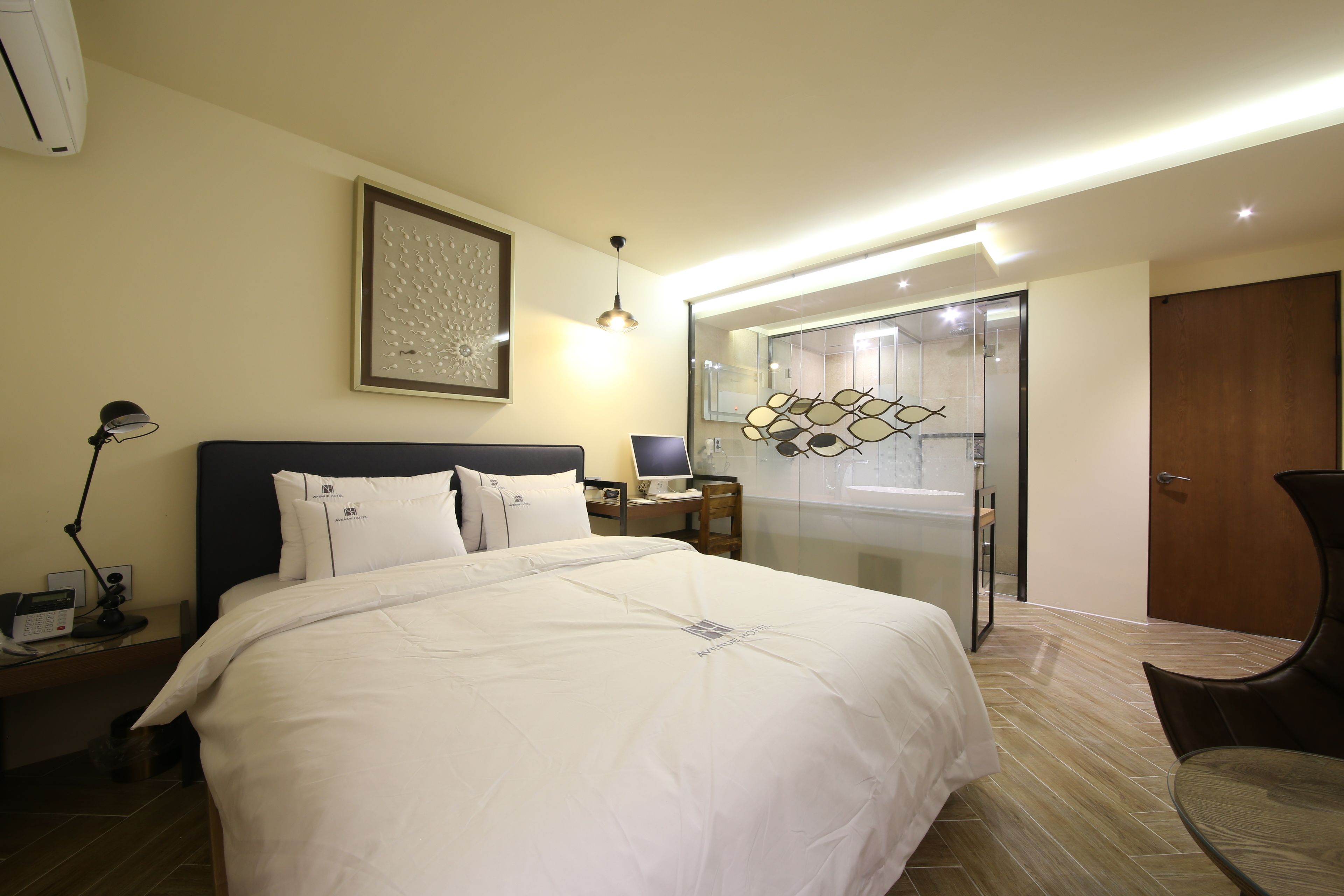 A hotel room with a large bed, bedside table with a lamp, a desk with a computer, and a glass shower enclosure.