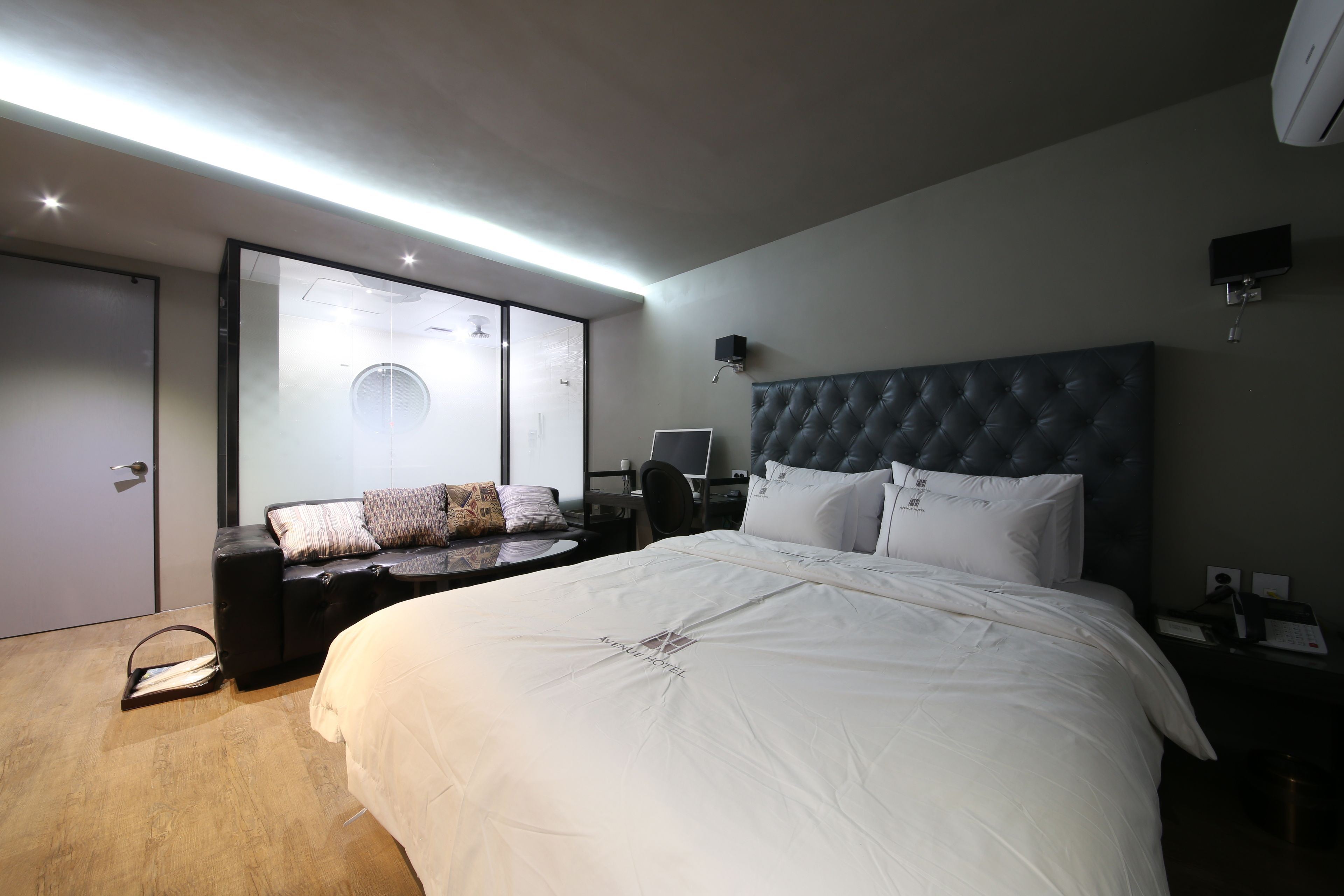 A modern hotel room with a large bed, a desk with a computer, a seating area with a couch, and a sliding glass door leading to a balcony.