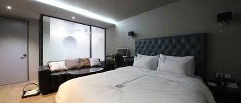 Una habitación de hotel moderna con una cama grande, un escritorio con computadora, una zona de estar con sofá y un ventanal corredizo que da a un balcón.