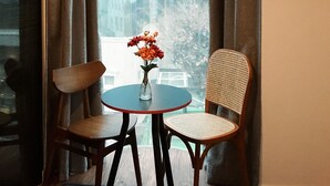 A room with a round table, two chairs, a vase with flowers, and a view of tall buildings through large windows. - ARA125 (Seoul)