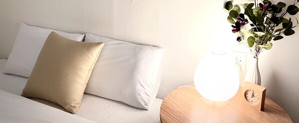 A neatly made bed with a beige pillow and a white pillow, a bedside table with a white lamp, a vase with flowers, and a wooden clock.