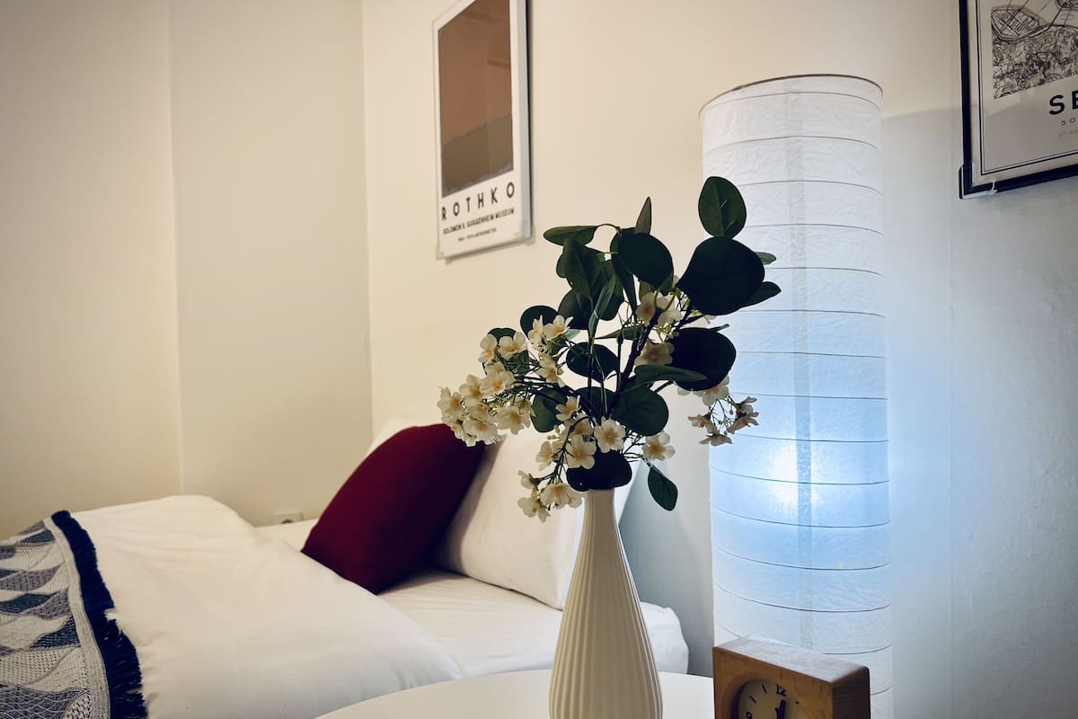 A white vase with flowers on a bedside table, a red pillow, a white bedspread, and a framed picture on the wall.