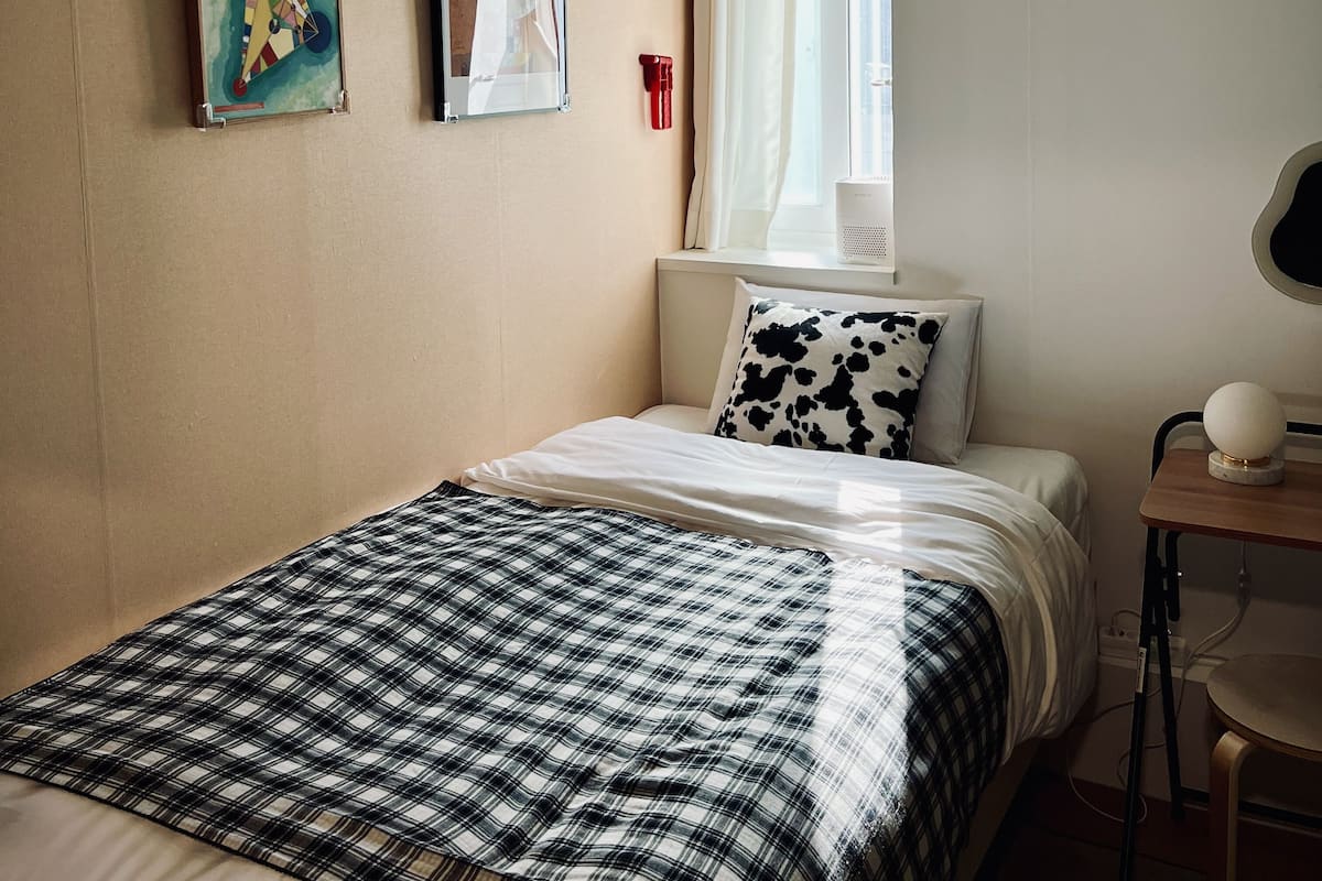 A bedroom with a bed, a window with curtains, a table with a lamp, and two framed artworks on the wall.