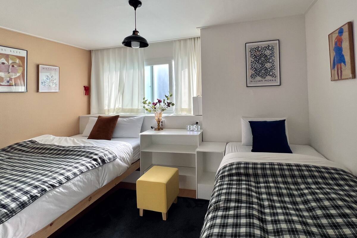 A bedroom with two beds, a small desk, a window with curtains, and wall art.
