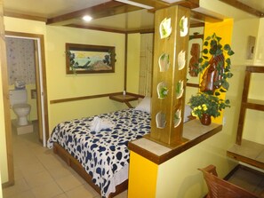 A room with a bed, a wooden headboard with decorative cutouts, a painting on the wall, a potted plant, and a toilet visible in the corner. - Cocopele Inn (San Ignacio)