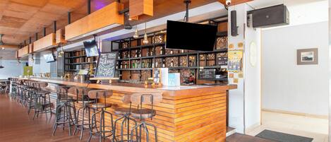 A bar area with wooden bar counter, stools, shelves with bottles, and a flat-screen TV mounted on the wall.