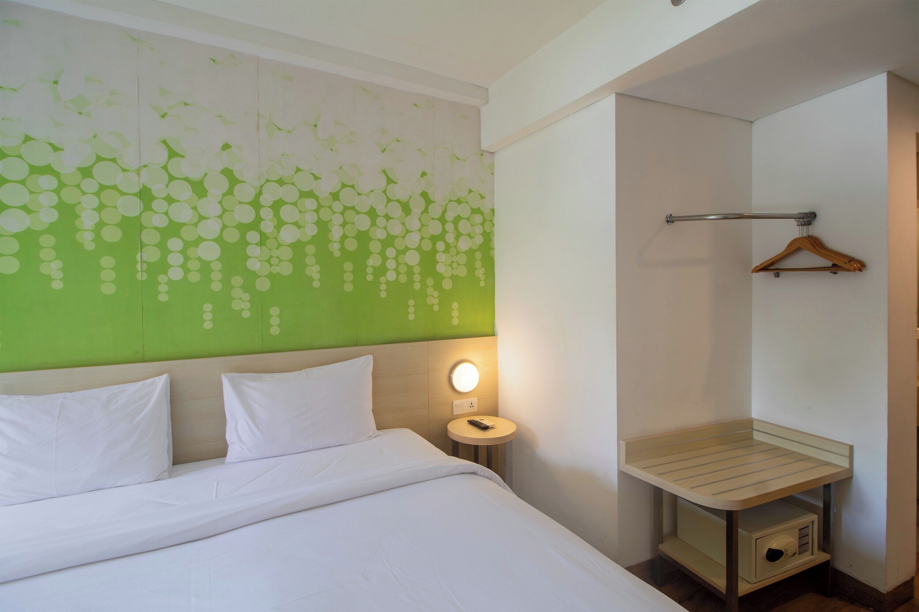 Une chambre d’hôtel comprenant un lit, une table de chevet avec une lampe, un porte-serviettes fixé au mur et une petite étagère en bois.