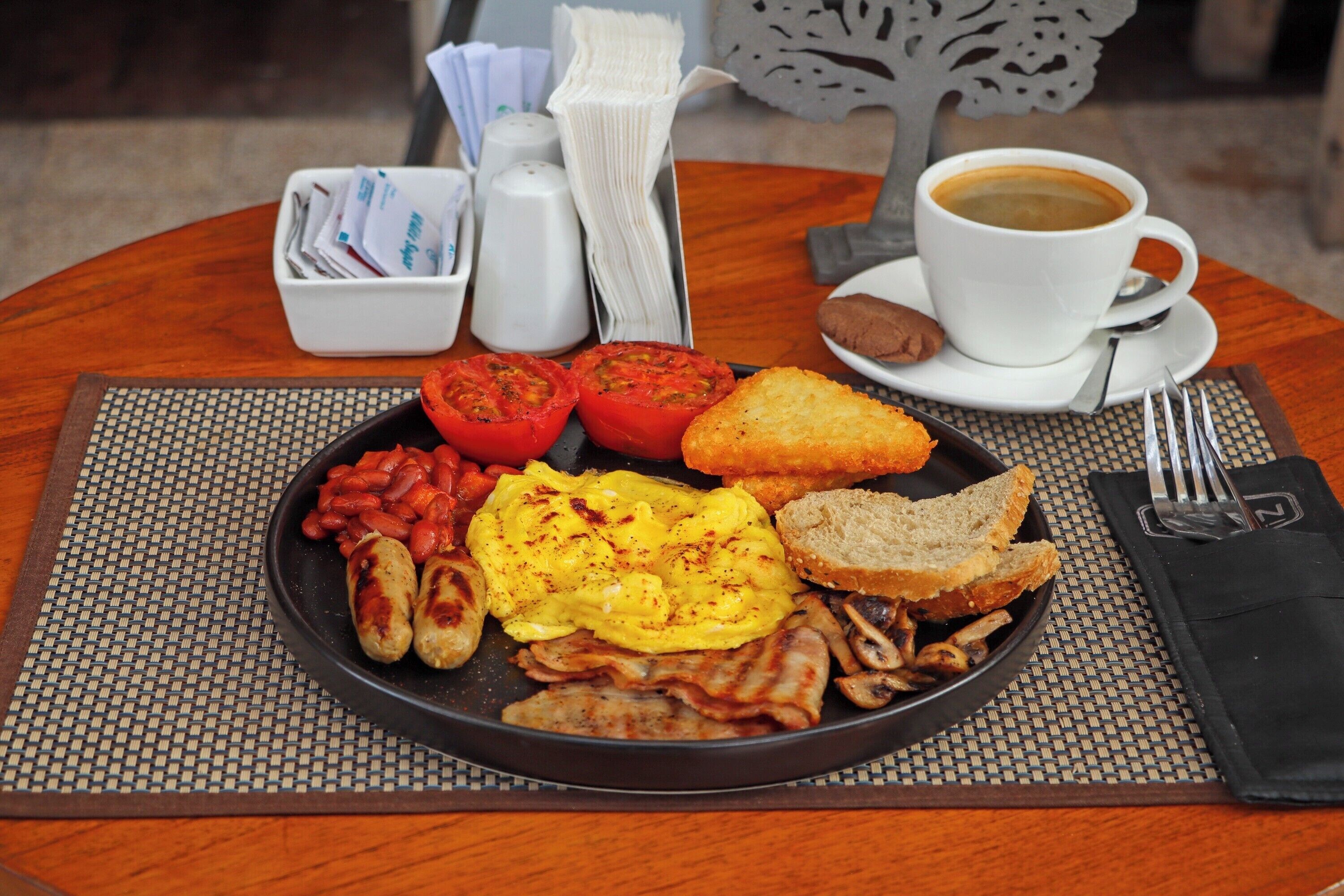 A breakfast plate with eggs, bacon, tomatoes, beans, toast, and a cup of coffee on a table.