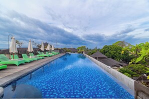 Piscina com espreguiçadeiras e guarda-sóis, cercada por vegetação e edifícios.