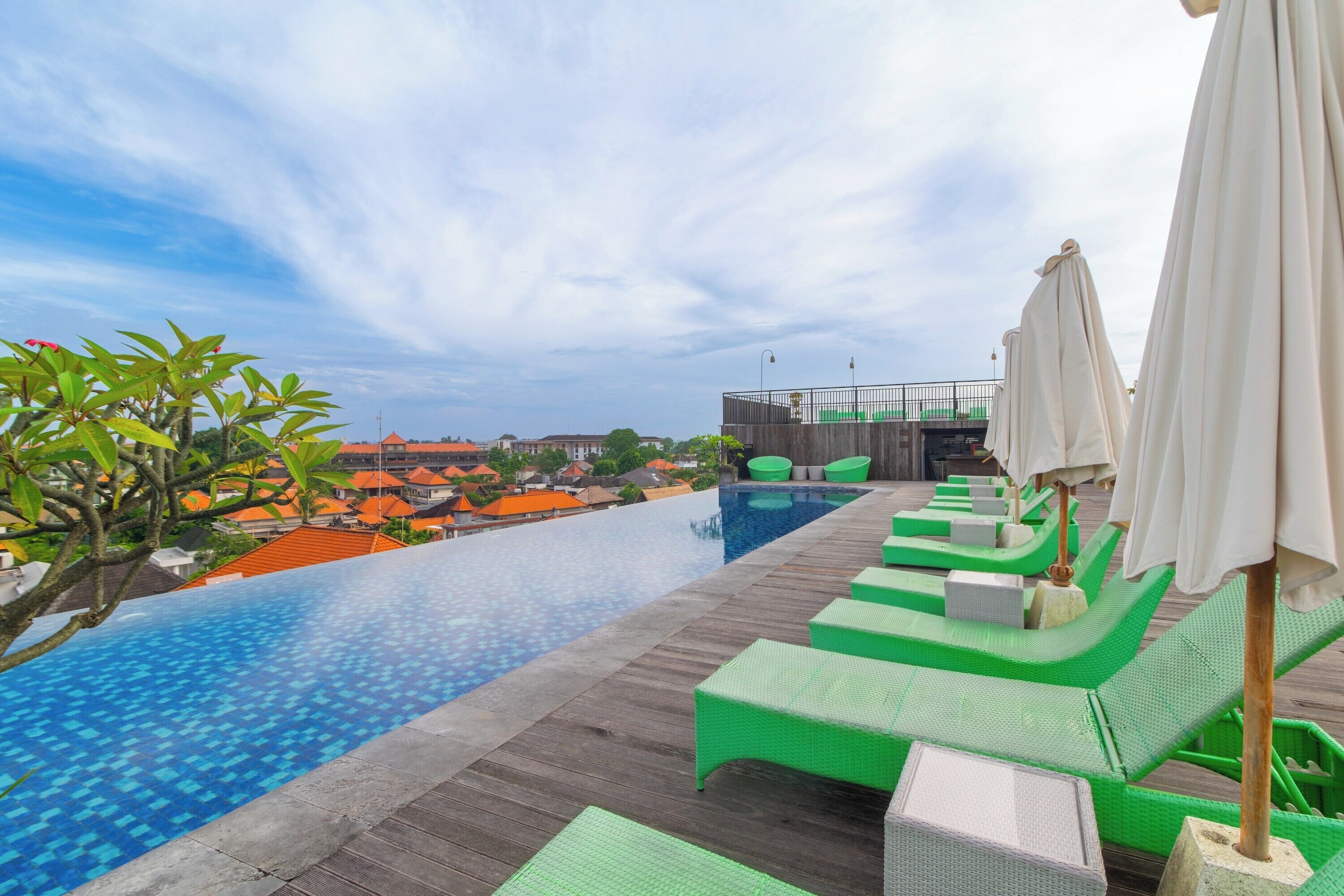 A rooftop infinity pool with green lounge chairs and umbrellas, overlooking a cityscape.