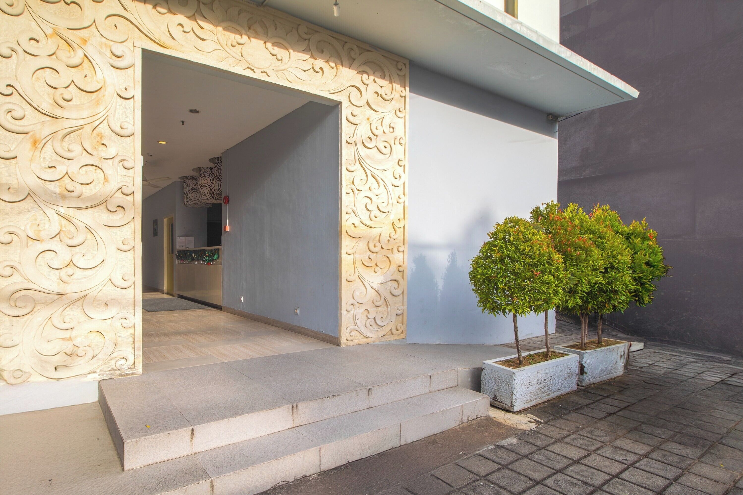 A building entrance with ornate wall carvings, a set of steps leading up to the door, and a small tree in a planter.